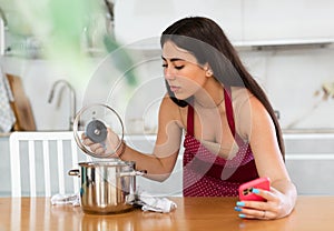 Young woman in apron with phone looks at pan