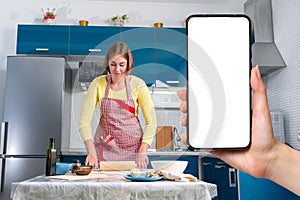A young woman in an apron rolls out the dough with a rolling pin. A woman& x27;s hand on the right holds a smartphone