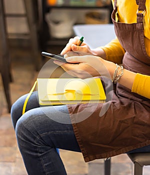 Young woman in apron working on smart phone, yellow paper notebook lies on her lap. Young woman using smart phone at class.