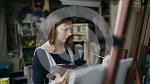 Young woman artist in apron painting picture on canvas in art studio