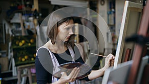 Young woman artist in apron painting picture on canvas in art studio