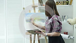 Young woman artist in apron painting picture on canvas in art studio