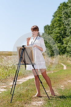 Young woman artist at the easel painting landscape outdoors. Girl-artist working on the plein air