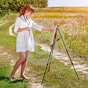 Young woman artist at the easel painting landscape outdoors. Girl-artist working on the plein air