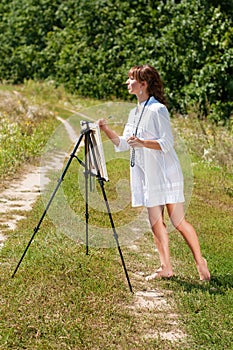 Young woman artist at the easel painting landscape outdoors. Girl-artist working on the plein air