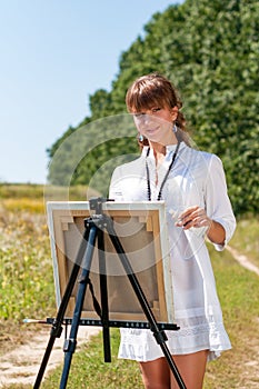 Young woman artist at the easel painting landscape outdoors. Girl-artist working on the plein air