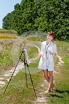 Young woman artist at the easel painting landscape outdoors. Girl-artist working on the plein air