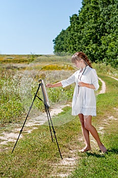 Young woman artist at the easel painting landscape outdoors. Girl-artist working on the plein air