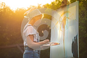 A young woman artist holds a brush and paints a picture on an easel in the rays of the sunset. The painter paints oil paintings in