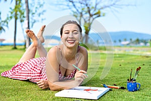 Young woman artist and illustrator drawing witha pencil, laying on a lawn in summer photo