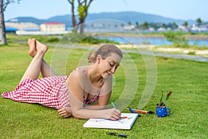 Young woman artist and illustrator drawing witha pencil, laying on a lawn in summer photo