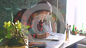 Young woman artist painting sketch on paper notebook with pencil. Bright sun flare from window