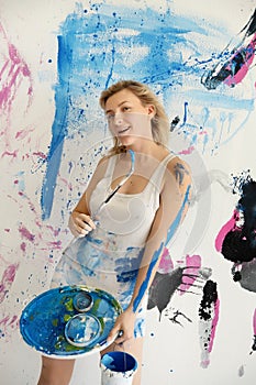 Young woman artist paints in color-stained white undershirt with blue paint, palette and brush with joy on the wall