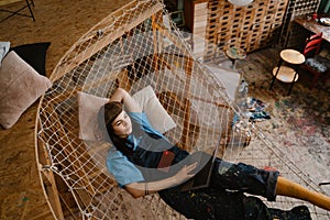 Young woman artist using laptop while resting in hammock at her studio
