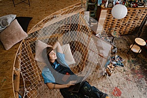 Young woman artist using laptop while resting in hammock at her studio