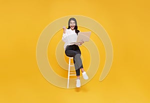 Young woman asian happy smiling. While her using laptop sitting on white chair and looking isolate on yellow background