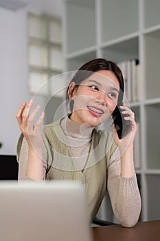 Young woman asian talking on mobile phone while working with laptop at desk home