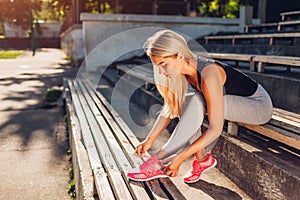 Young woman athlete tying sneakers sitting on bench on sportsground in summer. Preparation for training