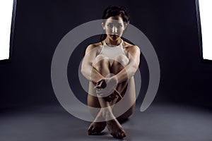 Young woman athlete in white top sitting on the floor between two studio flash lights embracing her crossed legs