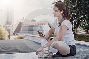 Young woman athletes in sportswear sitting in park, relax after sports training, use smartphone, listen to music.