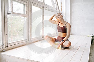Young woman with athletic body sitting in lotus position at window sill and stretching neck muscles