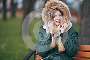 Young woman in autumn clothes sits on a bench in a city park. The woman is dressed in a stylish jacket with fur