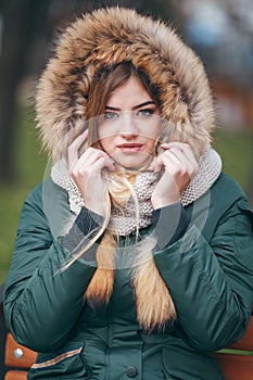 Young woman in autumn clothes sits on a bench in a city park. The woman is dressed in a stylish jacket with fur