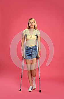 Young woman with axillary crutches on pink background