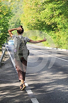 Young woman with backpack behind her back tries to stop passing car to hitchhike