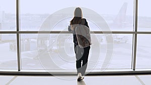 Young woman with backpack near terminal window. Caucasian female tourist using smartphone in airport lounge. Travel. 4K.