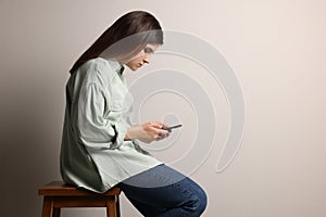 Young woman with bad posture using smartphone while sitting on stool against grey background. Space for text