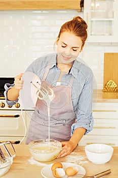 Young woman baking a cake in the kitchen standing at the counter in her apron using a handheld mixer