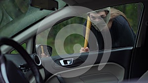 Young woman in balaclava with baseball bat looking inside car outdoors. Portrait of suspicious Caucasian lady robbing