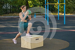 young woman with a ball in her hands makes lunges with her feet on a wooden box on a street sports ground.