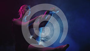 Young woman ballerina sits on the floor in black smoky studio and doing stretching