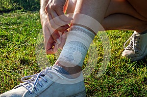 Young woman bandaging injured ankle. Injury leg while running outdoors. First aid for sprained ligament or tendon