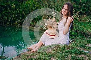 Young woman with bare feet sitting on the river bank. Brunette girl in a white dress near the lake with a bouquet of flowers