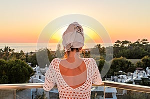 Young woman with a bath towel on her head stands on the balcony overlooking the sea at sunset time
