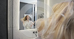 Young woman in bathrobe drying hair with a hairdryer in the bathroom at home. Usual morning beauty treatments in the