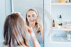 Young woman in bathrobe looking in the mirror, holding bottle with moisturizer cream and applying it on face. Skin