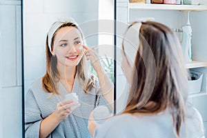 Young woman in bathrobe looking in the mirror, holding bottle with moisturizer cream and applying it on face. Skin