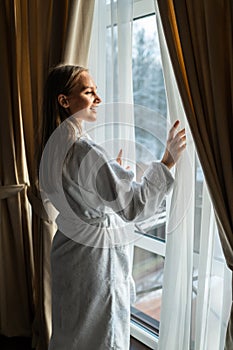 Young woman in bathrobe opening bedroom curtains at hotel room