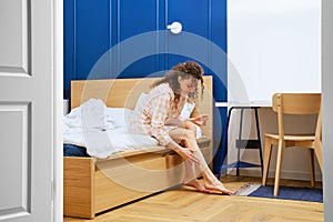 Young woman in bathrobe sitting on bed and applying moisturising cream