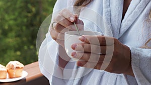 Young woman in bathrobe stirring sugar with spoon in cup of coffee while having breakfast on hotel room balcony or