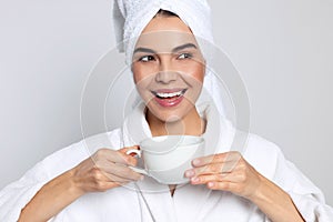 Young woman in bathrobe with towel and cup of drink on light grey background. Spa treatment