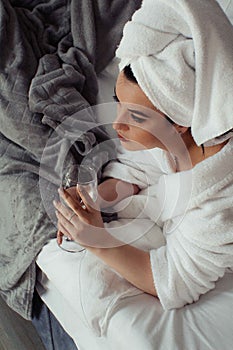 Young woman in a bathrobe and a towel on her head lies on the bed in a glass of water in her hand.