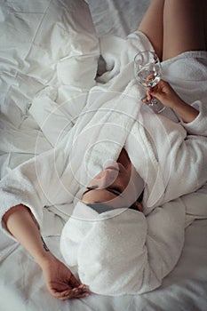 Young woman in a bathrobe and a towel on her head lies on the bed in a glass of water in her hand.