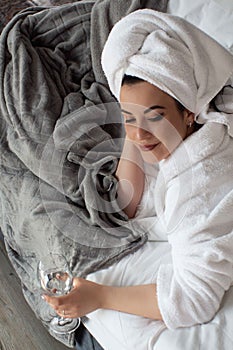 Young woman in a bathrobe and a towel on her head lies on the bed in a glass of water in her hand.