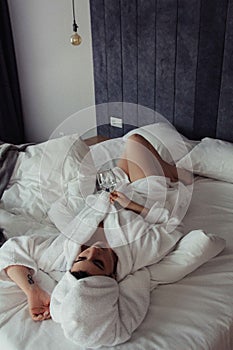 Young woman in a bathrobe and a towel on her head lies on the bed in a glass of water in her hand.