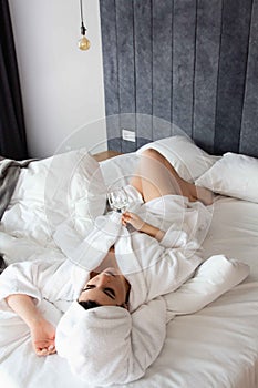 Young woman in a bathrobe and a towel on her head lies on the bed in a glass of water in her hand.